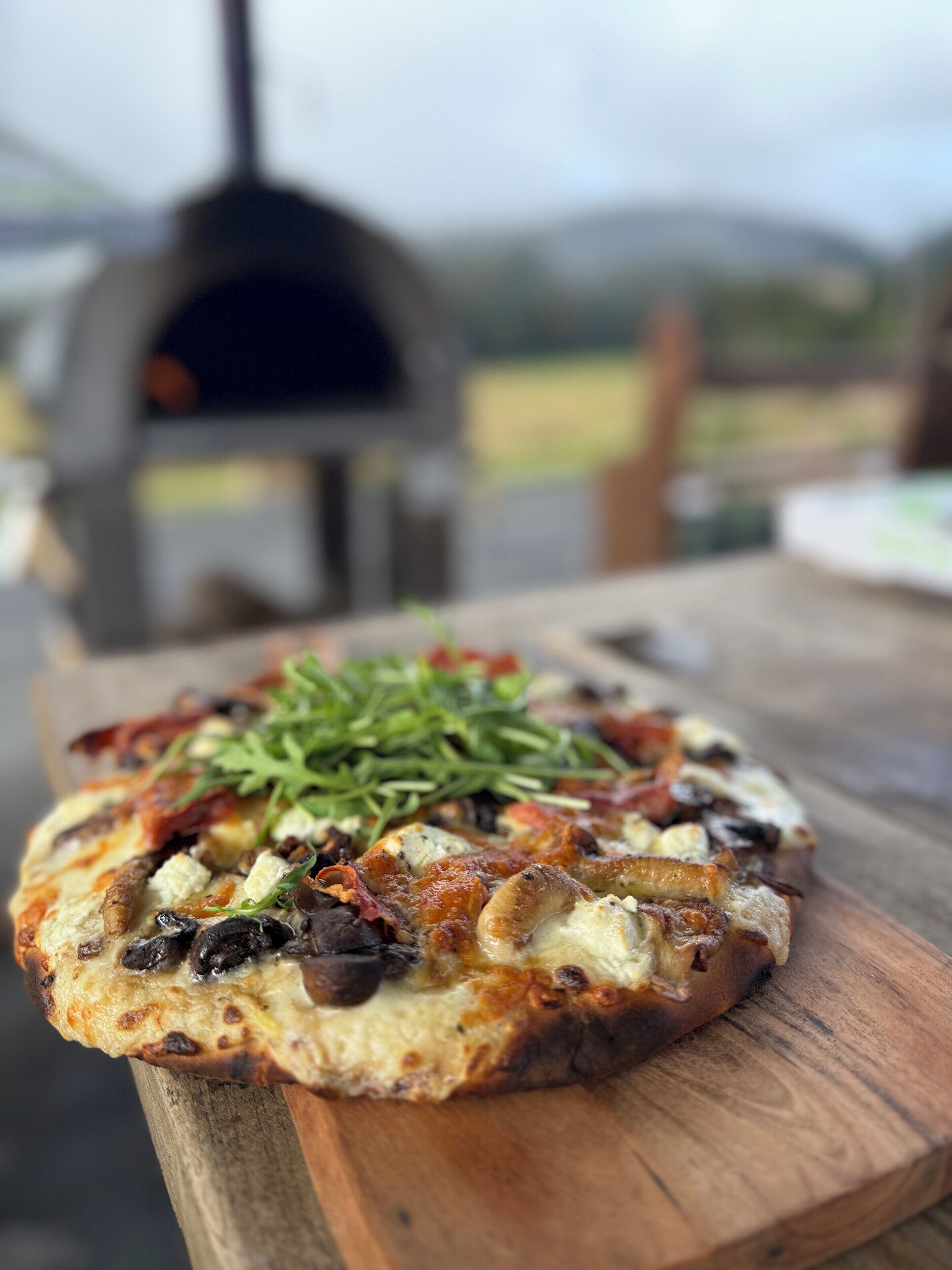 A pizza on a wooden board with a pizza oven in the background.