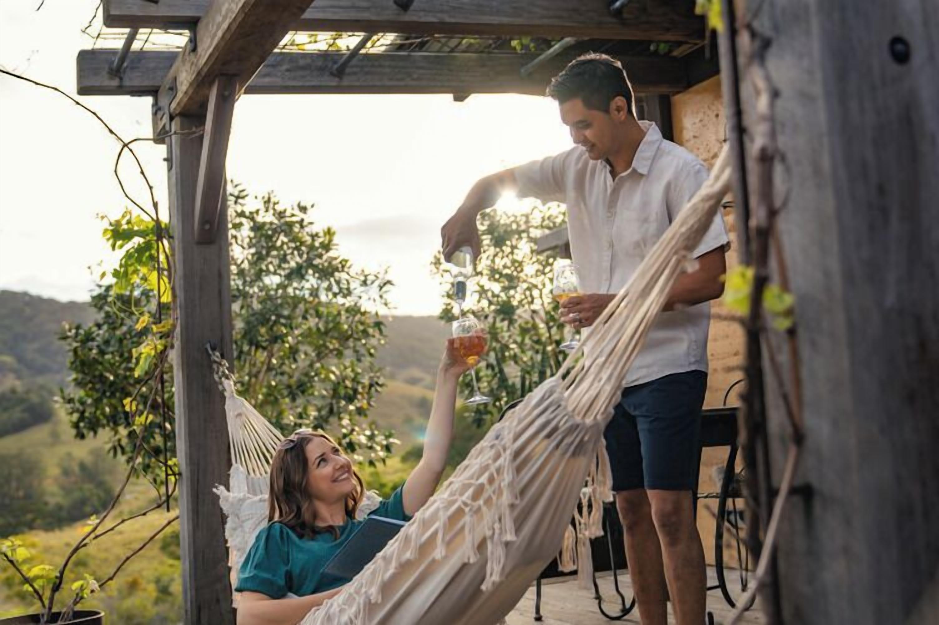 A hammock under a wooden pergola with a person standing and pouring a drink into a glass.