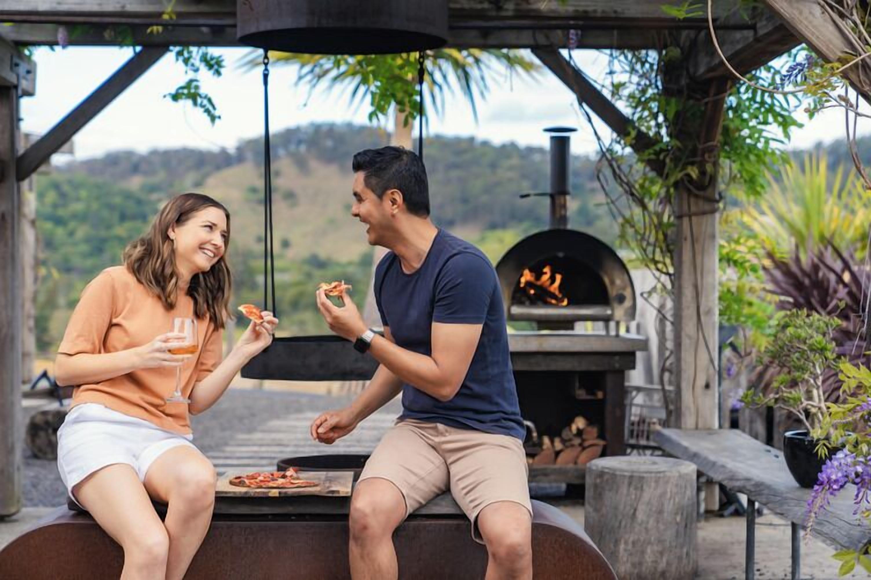 Deux personnes sont assises près d’un barbecue en plein air, une pizza à portée de main, entourées de verdure et avec une vue sur des collines.