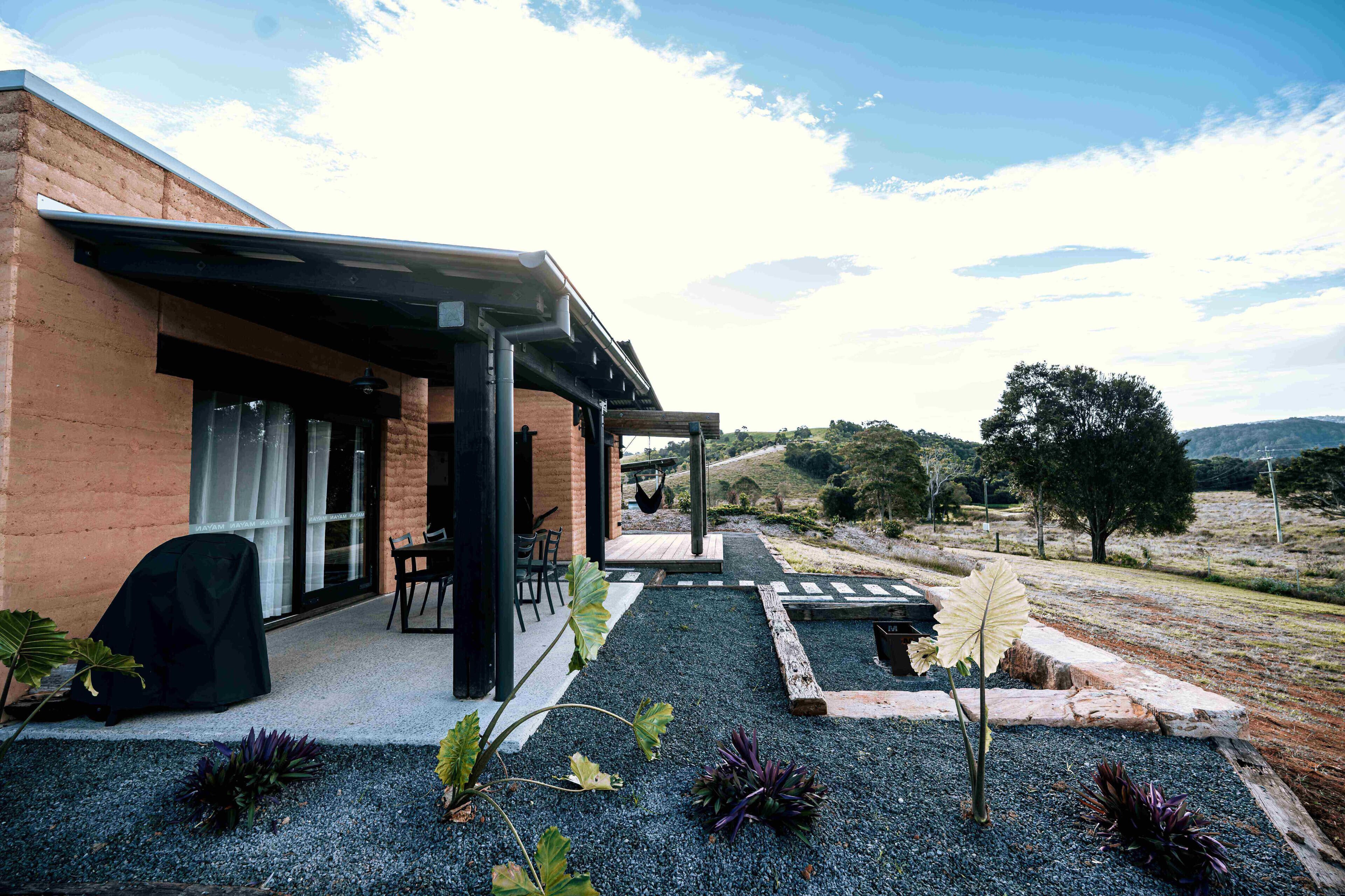 Une maison moderne dotée d’un patio couvert, d’un espace salon extérieur et d’un jardin agrémenté de gravier et de plantes.