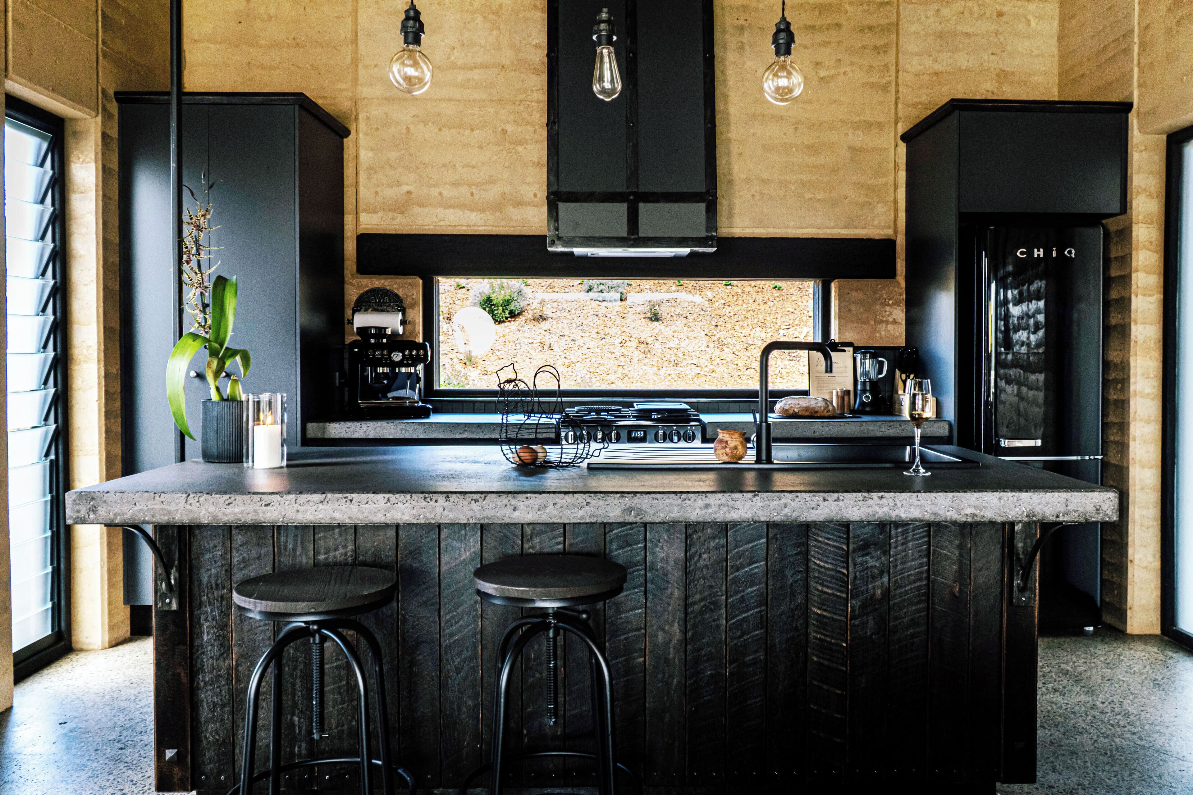 A modern kitchen with a central island, bar stools, a coffee machine, and a view of a garden through a window.