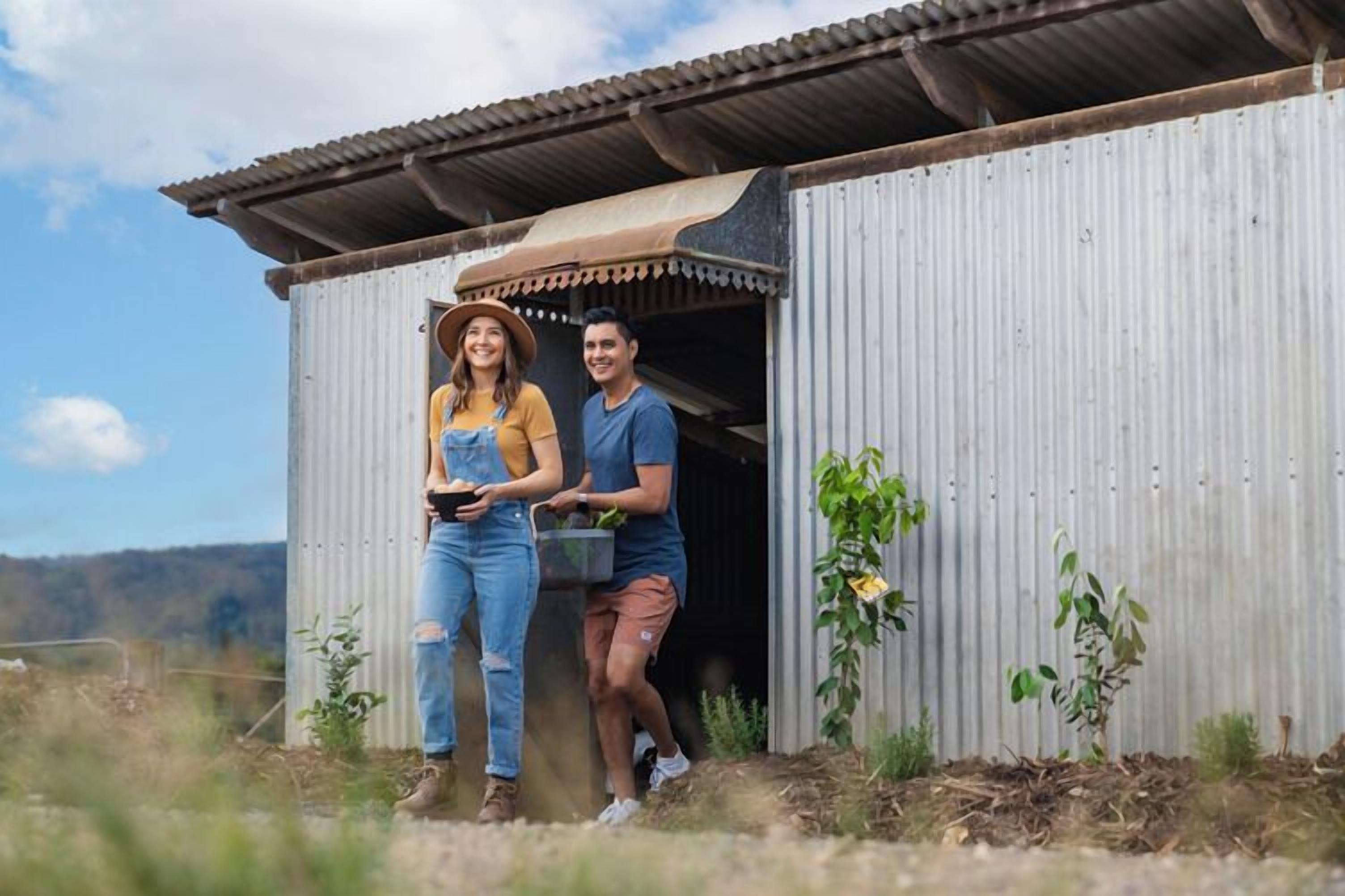 Two people standing outside a corrugated metal building with a brown roof, surrounded by small plants and a clear sky.
