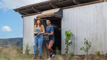 Two people standing outside a corrugated metal building with a brown roof, surrounded by small plants and a clear sky.