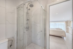 A modern bathroom with a glass-enclosed shower, marble walls, and a visible bedroom with a bed. - Mythos 2 (Agios Vasileios)