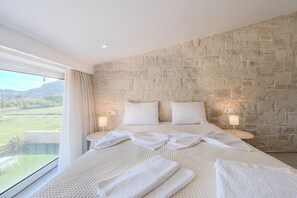 A bedroom with a stone wall, a large bed, two bedside lamps, and a view of a green landscape. - Mythos 3 (Agios Vasileios)
