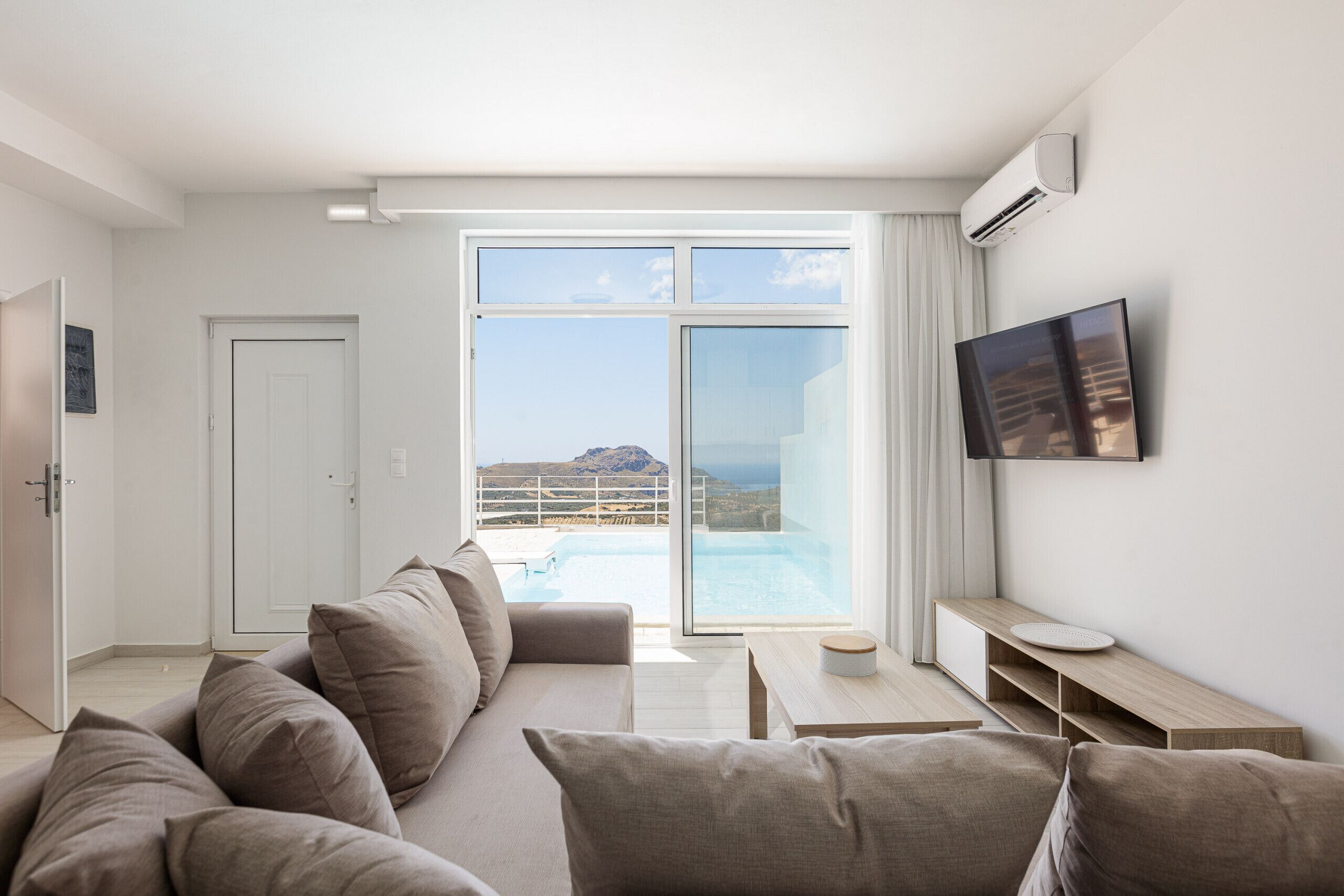 A modern living room with a sofa, a flat-screen TV, a small table, and a view of a pool and mountains.