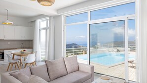 A modern living room with a large window overlooking a pool and a scenic view. - Petres Villas (Agios Vasileios)