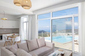 A modern living room with a large window overlooking a pool and a scenic view. - Petres Villas (Agios Vasileios)