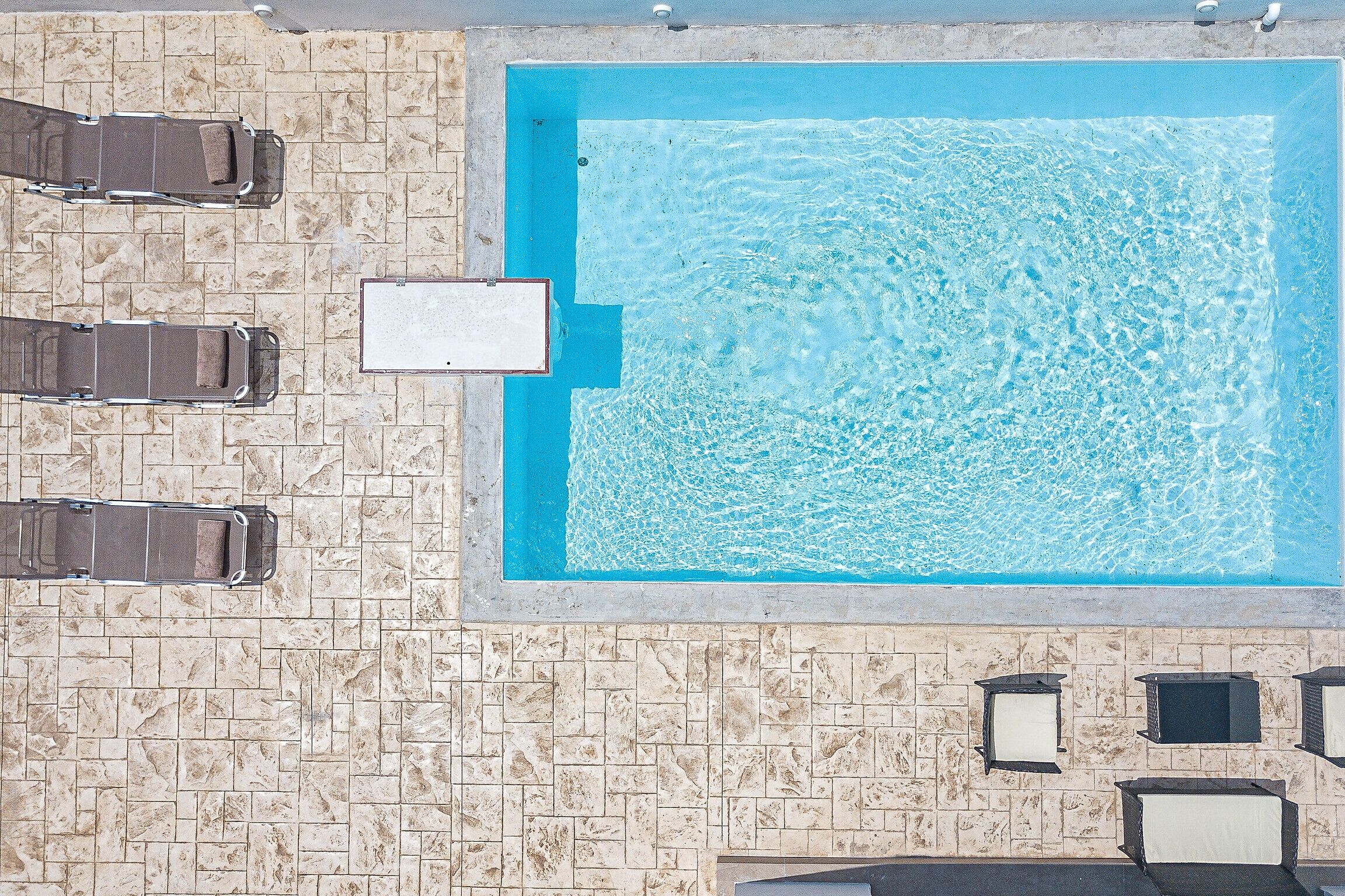 Vue aérienne d’une piscine rectangulaire dont le bord est recouvert de carreaux, entourée de chaises longues et d’une petite structure blanche.