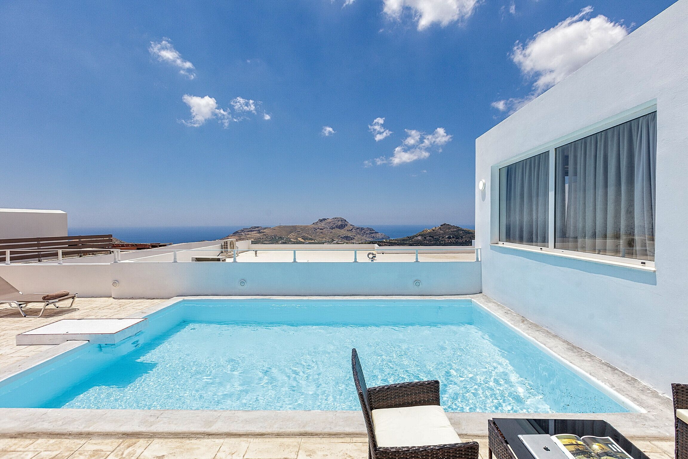 A rooftop pool with clear blue water, a view of the sea and mountains, and a white building with large windows.