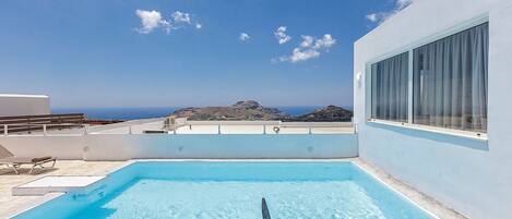 A rooftop pool with clear blue water, a view of the sea and mountains, and a white building with large windows.