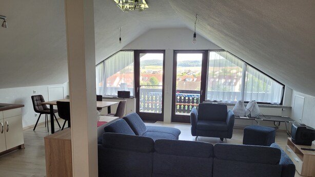 Una sala moderna con un sofá modular, una mesa de comedor con sillas, una zona de cocina y ventanas amplias que ofrecen vistas al exterior.
