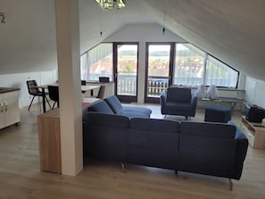 Ein modernes Wohnzimmer mit einer Eckcouch, einem Esstisch mit Stühlen, einer Küchenzeile und großen Fenstern mit Blick nach draußen.