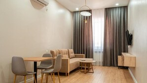 A modern living room with a wooden dining table, two grey chairs, a beige sofa, a small round coffee table, and a TV mounted on the wall. - BnB Hotel Shymkent (Shymkent)
