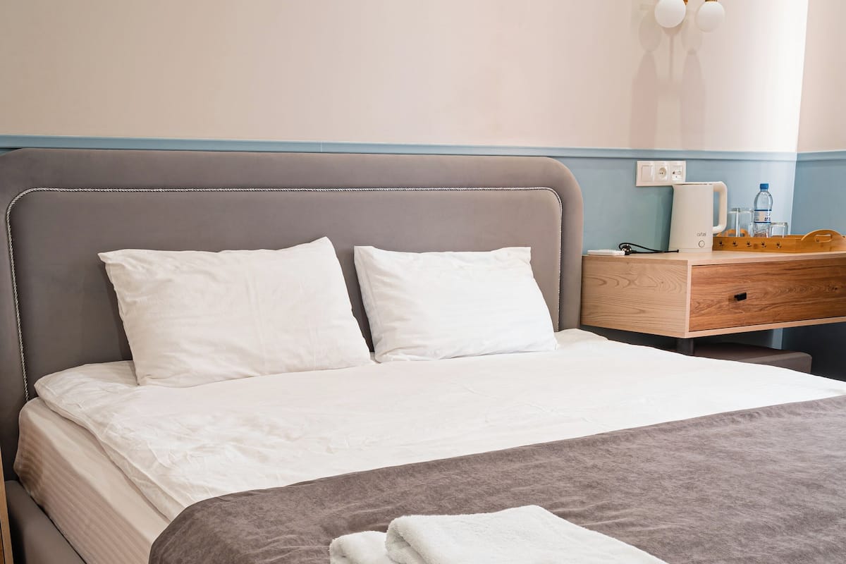 A neatly made bed with a grey headboard, a wooden nightstand with a lamp, and a wall-mounted light fixture.