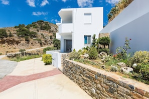 A modern white building with a stone retaining wall, a paved driveway, and a hillside with vegetation.