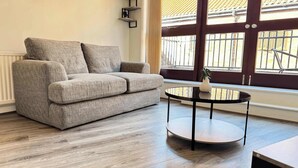 A living room with a grey sofa, a round black table, and a window with a view of a building with a tiled roof. - York City Centre Flat with view - Best Location - Walk to Everything (York)