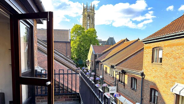 A balcony with a view of a church tower, brick buildings, and a street with umbrellas. - York City Centre Flat with view - Best Location - Walk to Everything (York)