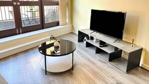 A modern living room with a round glass coffee table, a flat-screen TV on a black stand, and a large window with a view of outdoor furniture. - York City Centre Flat with view - Best Location - Walk to Everything (York)