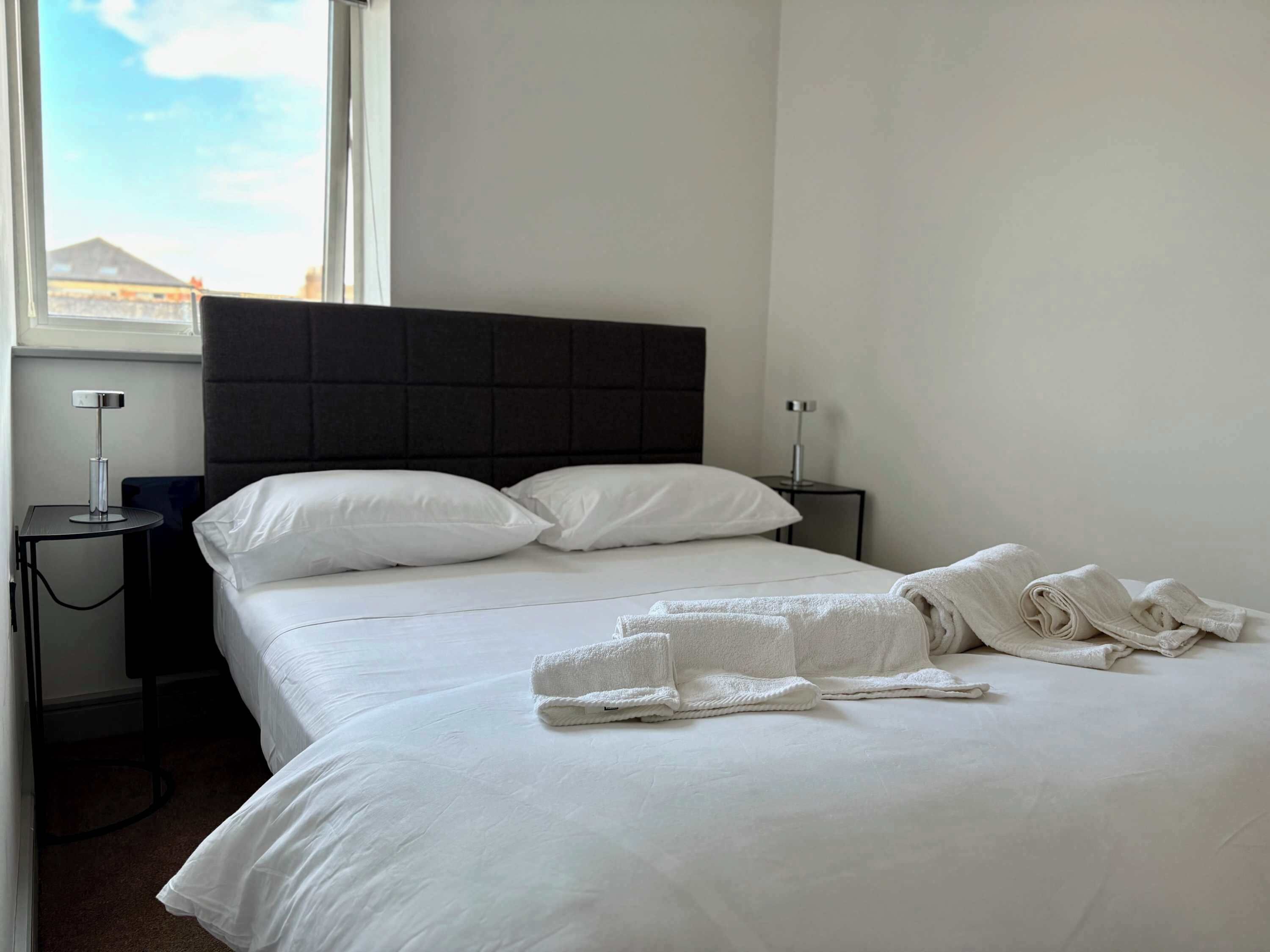 Hotelkamer met een bed, witte lakens, een donker hoofdeinde, twee nachtkastjes met lampen en een raam met uitzicht op gebouwen.