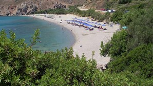 Een strand met een rij blauwe parasols, rotsachtige heuvels op de achtergrond en enkele gebouwen op de helling.