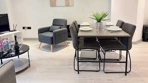 Un comedor moderno con una mesa dispuesta para cuatro, un sillón gris, una pequeña planta en maceta y un televisor sobre un mueble.