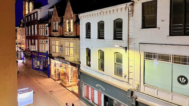 Vista de una calle en un barrio histórico urbano con arquitectura tradicional, incluyendo un edificio con el rótulo 'Rudy's' y otro con el rótulo 'Urban Outfitters'.