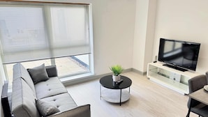 Una sala de estar moderna con un sofá gris, una mesa de centro redonda con una planta, un televisor de pantalla plana sobre un soporte blanco, y un comedor con una mesa y sillas.