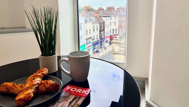 A table with croissants, a mug, and a York City Guide book, with a view of buildings through a window. - Modern 2-Bed Flat in York City Centre - Steps from Minster and Shambles (York)