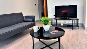 A modern living room with a grey sofa, a round black coffee table with a white mug and a magazine, a potted plant, and a TV displaying 'NETFLIX' on the screen. - Modern 2-Bed Flat in York City Centre - Steps from Minster and Shambles (York)