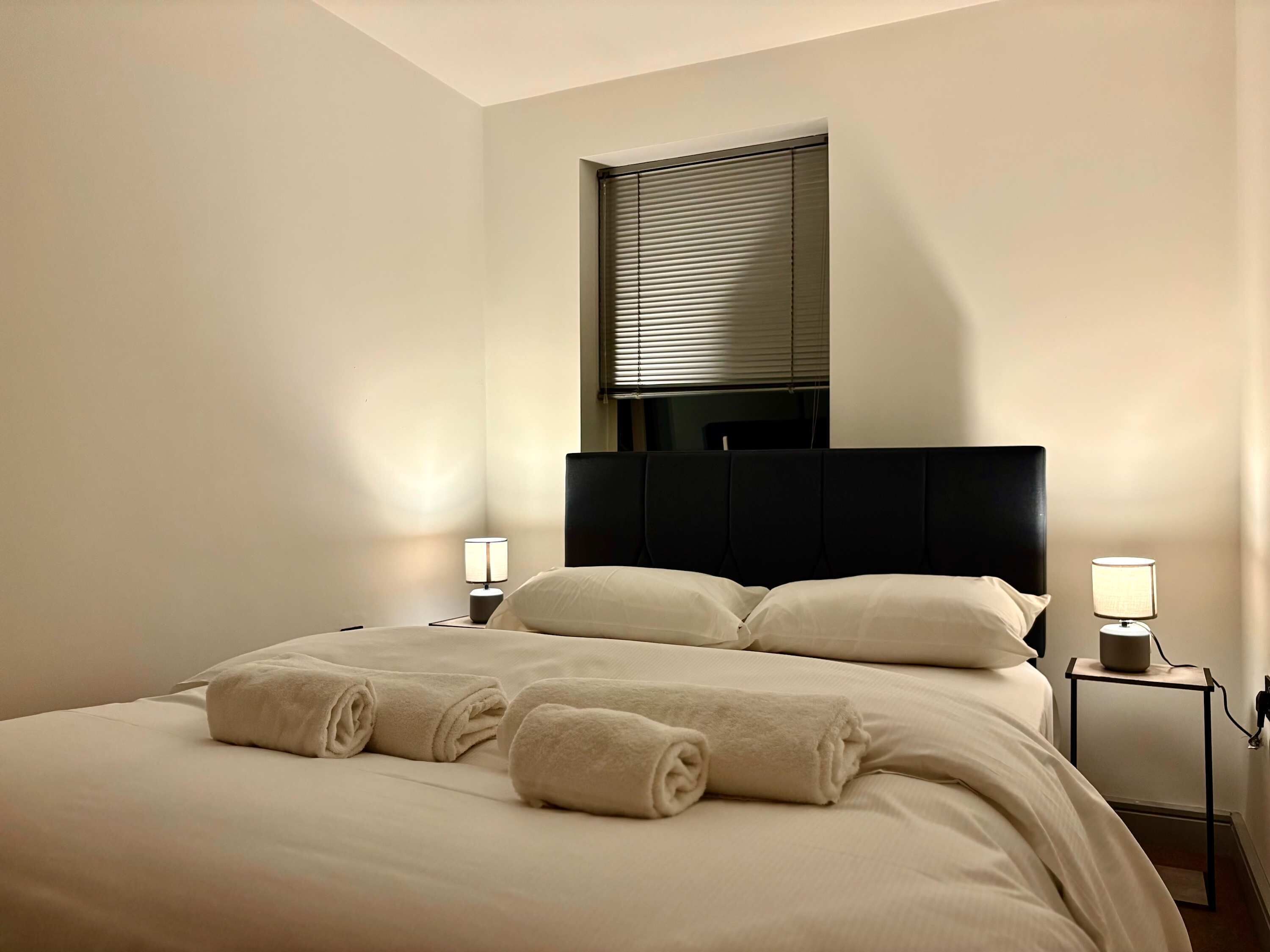 Habitación de hotel con una cama grande, dos mesitas de noche con lámparas, una ventana con persianas y un cabecero.
