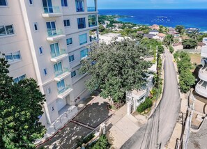 A multi-story building with balconies, a tree-lined street, and a view of the ocean.