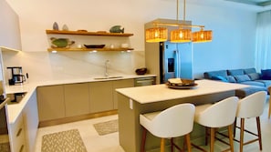A modern kitchen with a central island, bar stools, a coffee machine, and a dining area with a sofa and cushions.