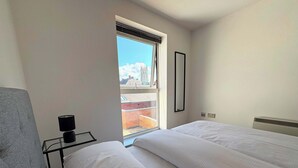 Un dormitorio con dos camas, una ventana que da a un edificio, una mesita de noche con una lámpara y un espejo montado en la pared.