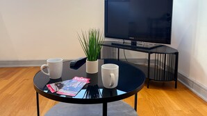 Una sala de estar moderna con un televisor negro sobre un mueble bajo, una pequeña mesa redonda con dos tazas blancas y una planta, y una revista sobre la mesa.