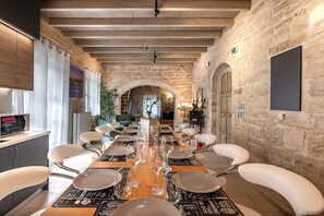 A dining area with a long table set for six, surrounded by white chairs, in a room with stone walls and wooden beams. - Alexandros 3 Villa (Rethymno)