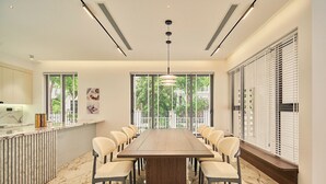 Een moderne eetruimte met een lange houten tafel, omringd door stoelen, grote ramen met zonwering en een keuken met een marmeren aanrecht.