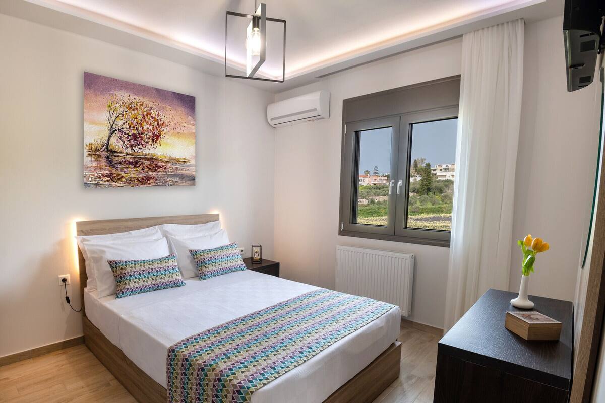 A modern bedroom with a large bed, a bedside table with a vase of flowers, a painting on the wall, and a window with a view of the outside.