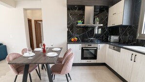 A modern kitchen with a marble backsplash, a built-in oven, and a dining area with pink chairs and a dark wooden table. - Destino Apartments (Rethymno)