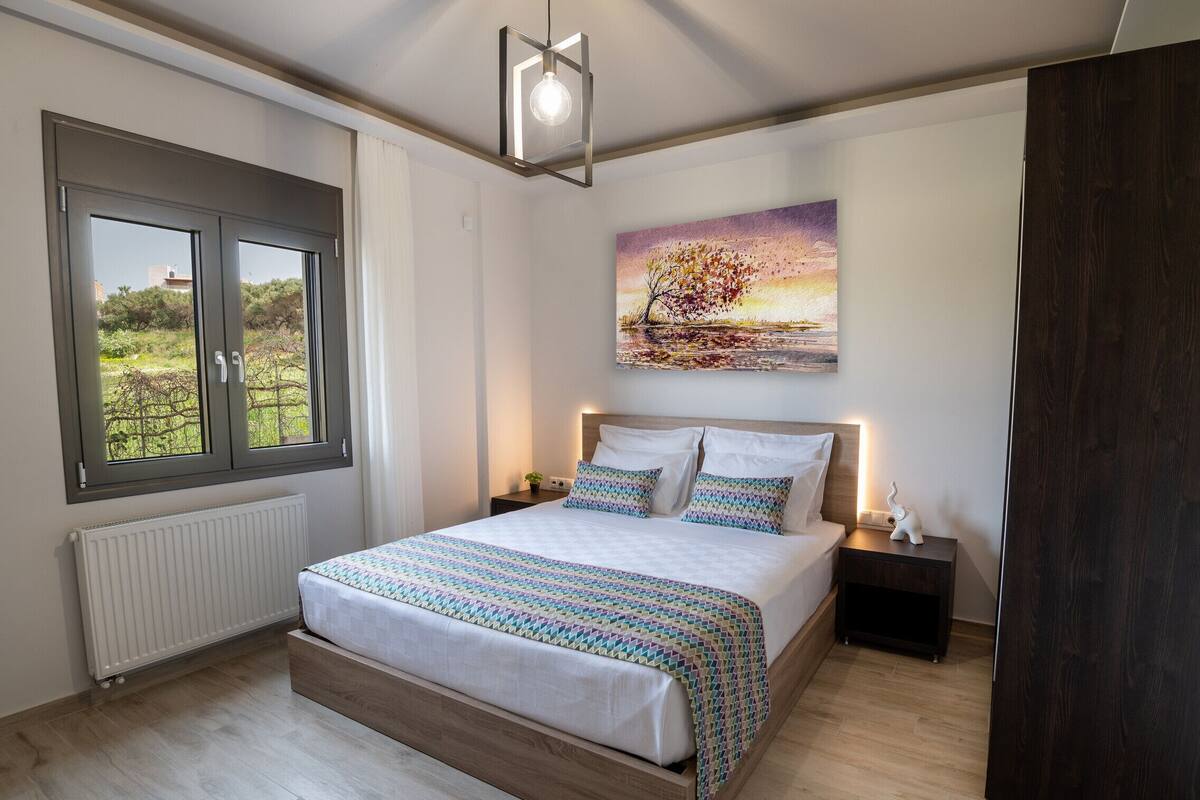 A bedroom with a large bed, a window with a view, a painting on the wall, and a bedside table with a lamp.