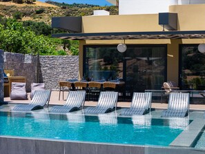 A modern outdoor pool area with lounge chairs, a dining table, and a stone wall. - Bella Vista Villa (Agios Vasileios)