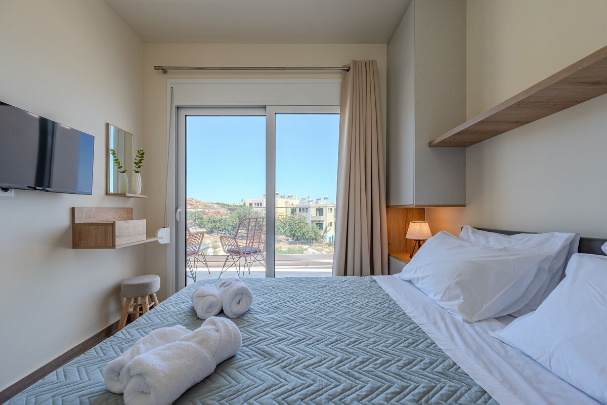 A hotel room with a bed, a TV, a chair, a window with curtains, and a view of buildings outside.
