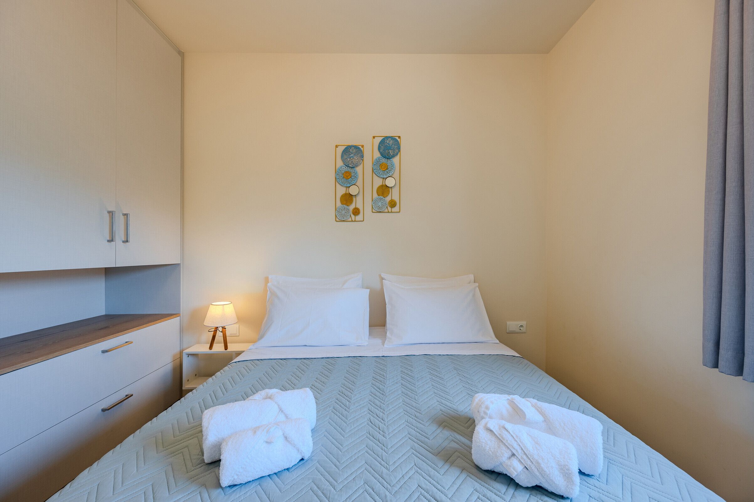 Een hotelkamer met een bed, twee kussens, een nachtkastje met een lamp, een garderobe en twee decoratieve wandobjecten.