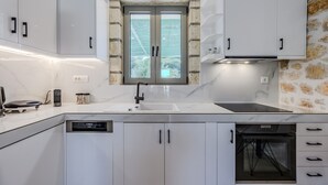 A modern kitchen with white cabinets, a built-in oven, and a window with a view of greenery. - Melidoni Villas (Mylopotamos)