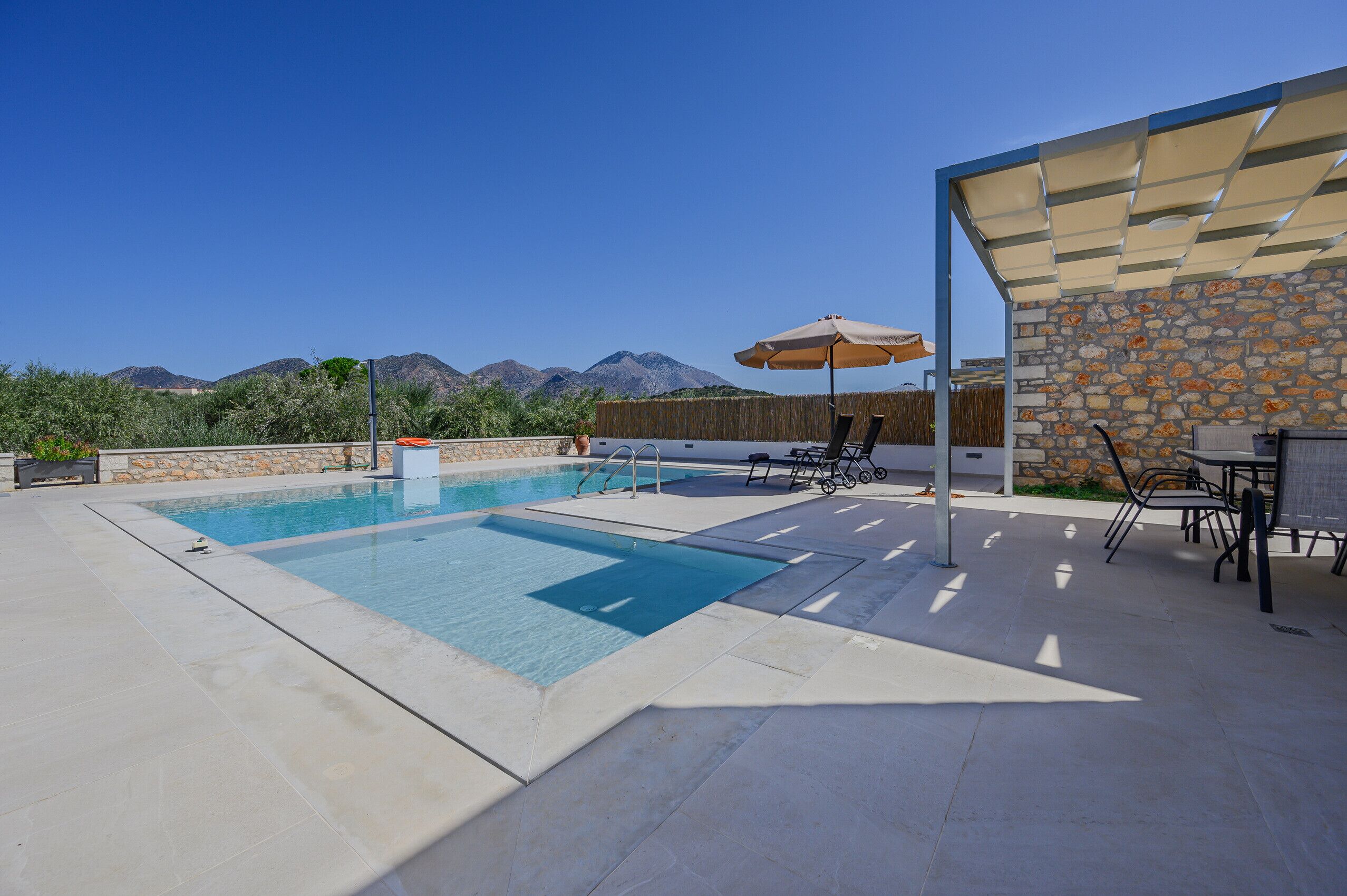 Ein Schwimmbecken mit einer Steinmauer und einer überdachten Terrasse mit Stühlen und Tischen.