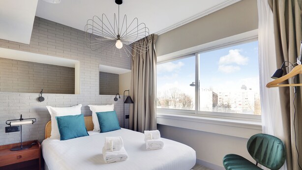 A modern hotel room with a large bed, a view of the city, and a green chair.