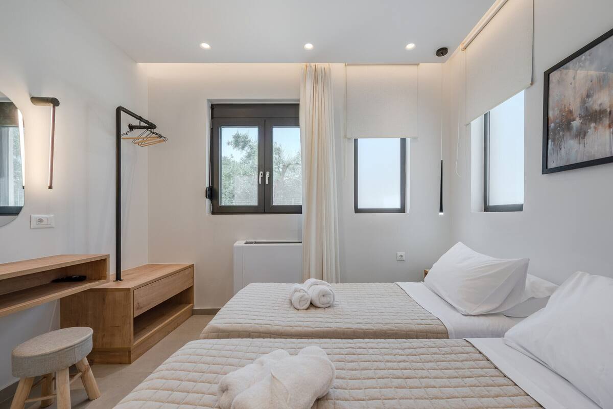 A modern bedroom with two beds, a wooden desk, a chair, and a large window.