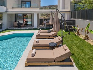 A modern outdoor pool area with lounge chairs and umbrellas, a dining area, and a view of a hillside. - Aether Group Villas (Rethymno)