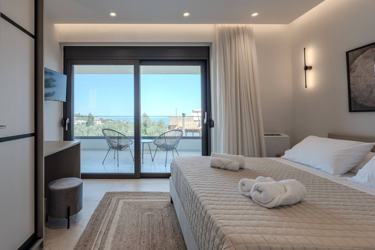 A modern hotel room with a large bed, a balcony with chairs, and a view of the city.
