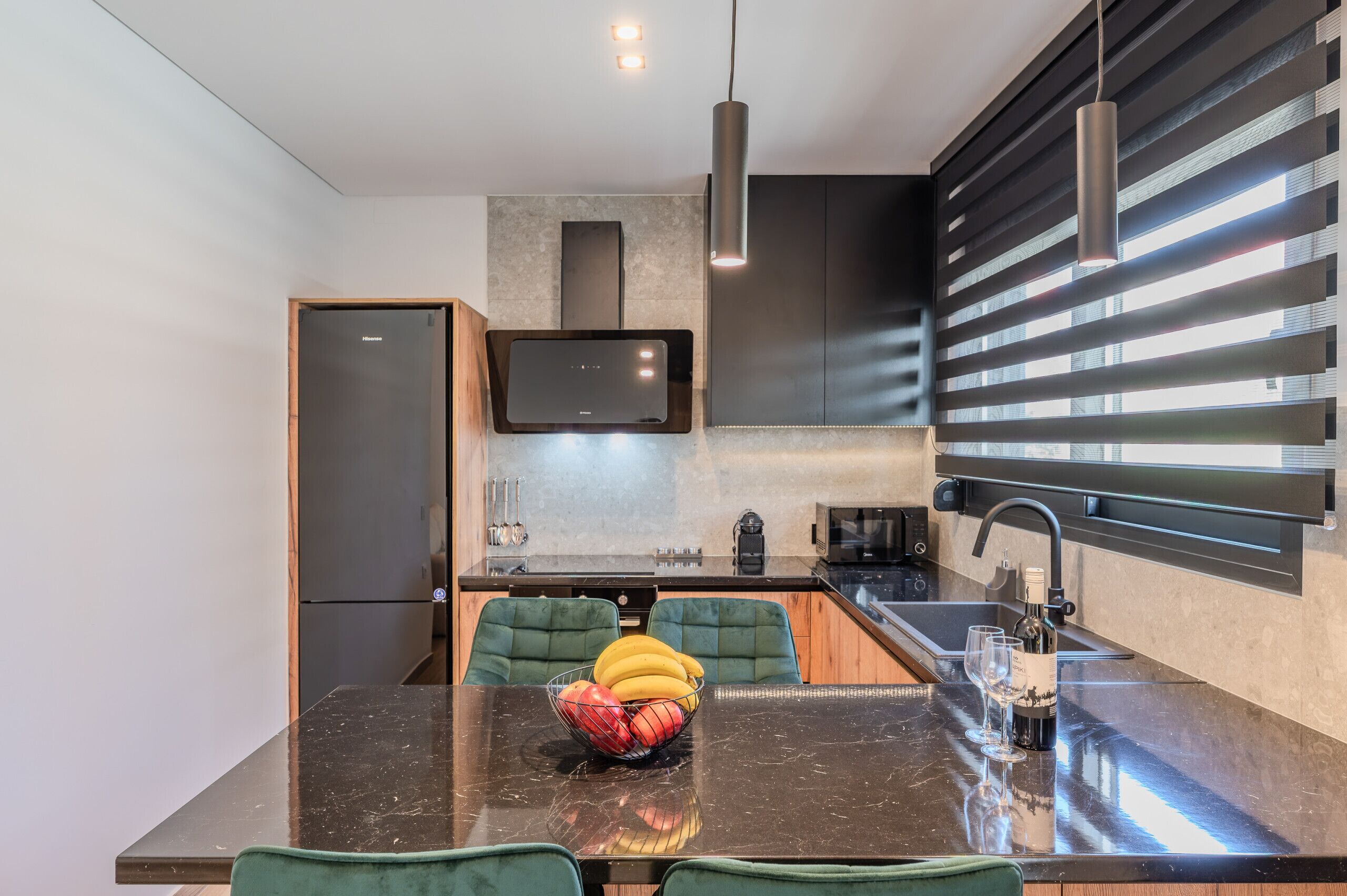 A modern kitchen with a black countertop, green upholstered chairs, a bowl of fruit, and a wine bottle.